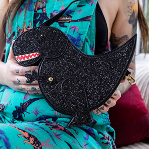 The kawaii black glitter dino vegan bag being held up by a tattooed model wearing dino print dungarees. The bag is facing forward to highlight the dinosaur face, moveable arm, detachable strap and black glitter front.
