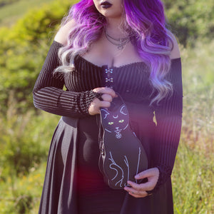 Model with purple hair wearing a goth outfit is holding the gothx pagan black cat vegan shoulder bag. The bag is facing forward to highlight the embroidery details including a pentagram collar, yellow eyes and paws.