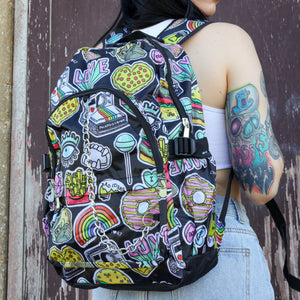 The kawaii graffiti doodle vegan backpack being worn by a tattooed model in front of a brown painted wall. The bag is facing forward to highlight the two front zip pockets, two elastic side pockets & silver chain. The all over print has lollipops, pugs, rainbows, pizzas, polaroid cameras, eyes and fries.