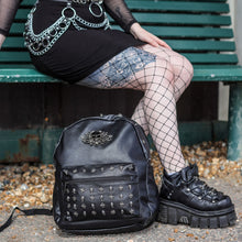 Load image into Gallery viewer, The GothX Skulls and Roses Vegan Backpack sat outside next to a gothic styled model with a black mini skirt, fishnets and chunky trainers on a bench. The black vegan leather backpack has silver skull and cross studs along the front zip pocket and along the top zip compartment along with a rose skull emblem in the middle.
