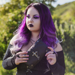 A model with long purple gradient hair in a goth outfit holding the gothx don't cross me vegan clutch bag. The clutch is facing forward to highlight the cross studs and stud, chain cross centrepiece.