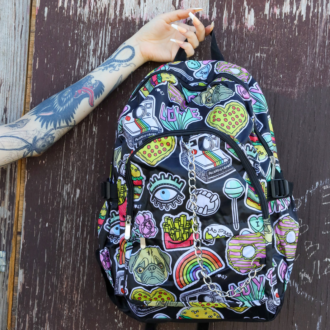 The kawaii graffiti doodle vegan backpack being held up in front of a brown painted wall by a tattooed model. The bag is facing forward to highlight the two front zip pockets, two elastic side pockets and silver chain. The all over print has lollipops, pugs, rainbows, pizzas, polaroid cameras, eyes and fries.
