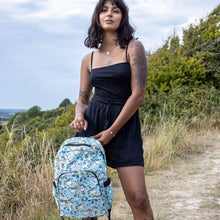 Load image into Gallery viewer, Meera is stood in a grass field wearing the kawaii unicorn vegan backpack on her back. The backpack is facing the camera, highlighting the pastel blue rainbow unicorn print, two front zip pockets, two side pockets and detachable silver chain.

