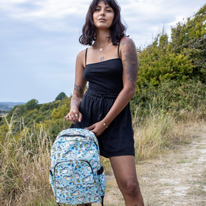 Meera is stood in a grass field wearing the kawaii unicorn vegan backpack on her back. The backpack is facing the camera, highlighting the pastel blue rainbow unicorn print, two front zip pockets, two side pockets and detachable silver chain.