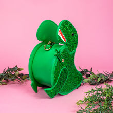 Load image into Gallery viewer, The kawaii green glitter dino vegan bag on a pink studio background with green foliage. The bag is facing forward angled left to highlight the green glittered side, dinosaur face, gold metal detailing, detachable strap and moveable arm.
