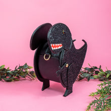 Load image into Gallery viewer, The kawaii black glitter dino vegan bag on a pink studio background with green foliage surrounding it. The bag is angled left and forward to highlight the dinosaur face, moveable arm, detachable strap and black glitter front.
