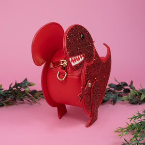 The kawaii red glitter dino vegan bag on a pink studio background with green foliage. The vegan leather red sparkly bag is facing forward angled left to highlight the red glittered side, dinosaur face, gold metal detailing, detachable strap and moveable arm.