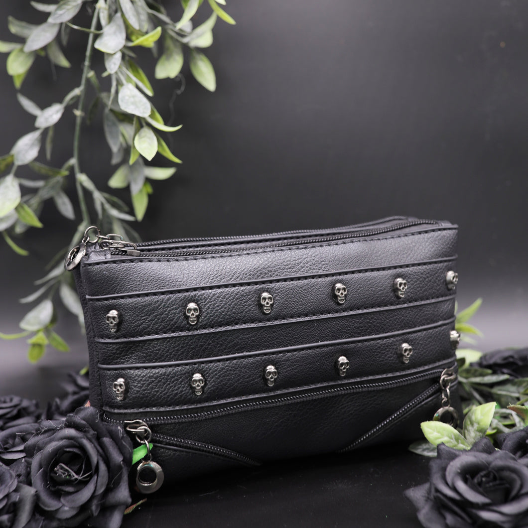The gothx mini skull head vegan clutch bag on a black studio background with black roses and green leaf foliage surrounding it. The clutch is facing forward to highlight the skull studded front and faux zip detailing.