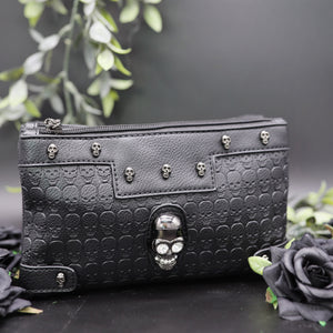 The gothx crystal skull vegan clutch bag on a black studio background with black roses and green foliage surrounding it. The bag is facing forward to highlight the embossed skull vegan black leather, mini skull studs, two main zip compartments and crystal skull centrepiece.