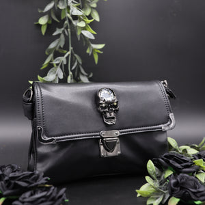 The GothX crystal skull vegan shoulder bag on a black studio background with black roses and green foliage surrounding it. The bag is facing forward to highlight the metal clasp close, metal trim detailing and crystal skull centrepiece.