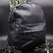 Load image into Gallery viewer, The GothX embossed skull vegan backpack on a black studio background with black roses and green foliage surrounding it. The bag is facing forward angled right to highlight the all over embossed skull design, front zip pocket and studded main zip compartment.
