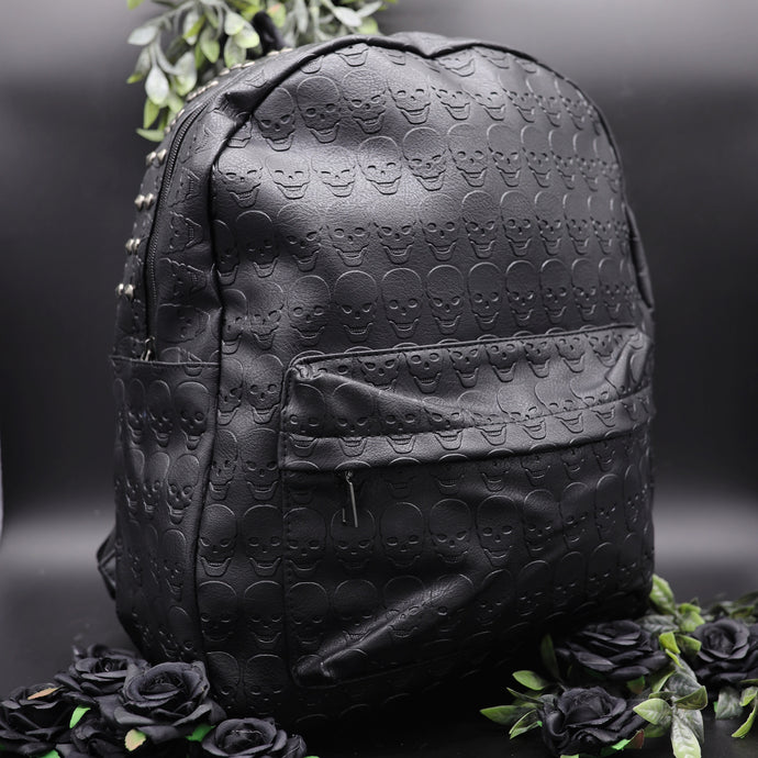 The GothX embossed skull vegan backpack on a black studio background with black roses and green foliage surrounding it. The bag is facing forward angled right to highlight the all over embossed skull design, front zip pocket and studded main zip compartment.