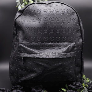 The GothX embossed skull vegan backpack on a black studio background with black roses and green foliage surrounding it. The bag is facing forward to highlight the all over embossed skull design, front zip pocket and studded main zip compartment.