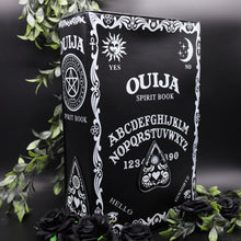 Load image into Gallery viewer, The Gothx ouija spirit book vegan backpack on a black studio background surrounded by black roses and green foliage. The bag is facing forward angled slightly right to highlight the embroidered planchette and white printed detailing featuring a ouija board, pentagrams and lace pattern.

