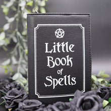 Load image into Gallery viewer, The gothx little book of spells vegan shoulder bag sat on a black studio background with green foliage and black roses surrounding it. The witchy inspired vegan leather bag is facing towards the camera to highlight the white printed design of a pentagram, framing and text reading little book of spells.
