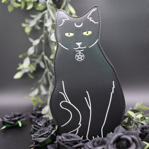 The gothx pagan black cat vegan shoulder bag on a black studio background with green foliage and black roses surrounding it. The bag is facing forward to highlight the embroidery details including a pentagram collar, yellow eyes and paws. 