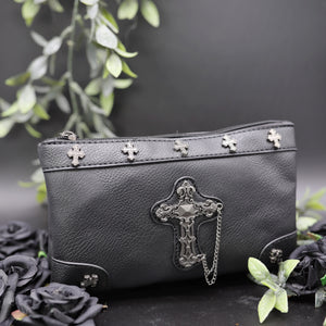 The gothx don't cross me vegan clutch bag on a black studio background with black roses and green foliage surrounding it. The clutch is facing forward to highlight the cross studs and stud, chain cross centrepiece.
