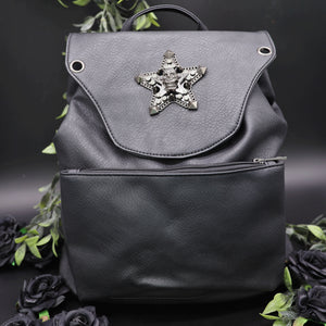 The GothX Skull and Star Black Vegan Tassel Tie Backpack in front of a black studio background with black decorative roses and leaves surrounding it. Black mini backpack bag with metal stud star with chains, faux crystals and skull head in the middle. Metal magnetic clip close and zip up large front pocket. Facing forwards to show off the detailing on the vegan leather bag.