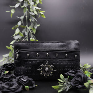 The gothx black skull vegan clutch bag on a black studio background with black roses and green foliage surrounding it. The clutch is facing forward to highlight the skull embossed vegan black leather, the crystal stud skull centrepiece, zip pocket and skull studs.