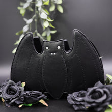 Load image into Gallery viewer, The GothX Black Bat Vegan Shoulder Bag on a black background with black roses and green foliage surrounding it. The bag is facing forward to highlight the embroidered detailing and crystal effect eyes.
