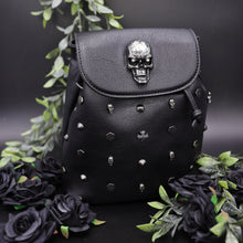 Load image into Gallery viewer, The GothX crystal skull vegan tassel tie backpack on a black studio background with black roses and green foliage surrounding it. The bag is facing forward to highlight the crystal skull magnetic close clip flap, tassel tie cords and cross, hexagon, cone and skull studs.
