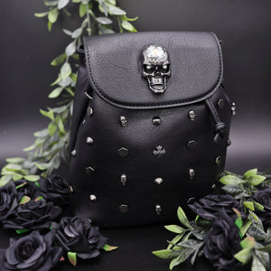 The GothX crystal skull vegan tassel tie backpack on a black studio background with black roses and green foliage surrounding it. The bag is facing forward to highlight the crystal skull magnetic close clip flap, tassel tie cords and cross, hexagon, cone and skull studs.