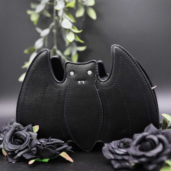 The GothX Black Bat Vegan Shoulder Bag on a black background with black roses and green foliage surrounding it. The bag is facing forward to highlight the embroidered detailing and crystal effect eyes.