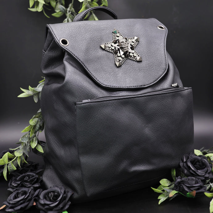 The GothX Skull and Star Black Vegan Tassel Tie Backpack in front of a black studio background with black decorative roses and leaves surrounding it. Black mini backpack bag with metal stud star with chains, faux crystals and skull head in the middle. Metal magnetic clip close and zip up large front pocket. Angled towards the right to show off the depth of the vegan leather bag.