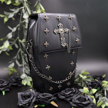 Load image into Gallery viewer, The GothX mini coffin vegan cross body bag. A 3d coffin shape bag on a black background with black roses and green foliage surrounding it. The vegan leather bag is facing forward angled to the right to highlight the detachable chain, cross studded front and stud cross centrepiece with chain.
