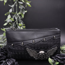 Load image into Gallery viewer, The gothx winged skull vegan clutch bag on a black studio background with black roses and green foliage surrounding it. The bag is facing forward to highlight the embossed skull leather, mini skull studs and winged skull centrepiece. 
