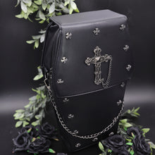 Load image into Gallery viewer, The gothx coffin vegan backpack is sat on a black studio background surrounded by black roses and green foliage. The bag is facing forward to highlight the cross mini studs, cross and chain emblem and detachable silver chain.
