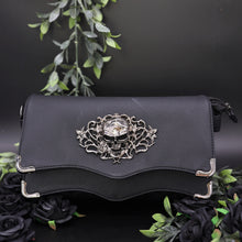 Load image into Gallery viewer, The GothX Skulls and Roses Quilted Clutch Bag sat on a black background with green foliage and black roses surrounding it. The vegan leather clutch bag is facing forward to highlight the two magnetic clip close flaps with metal corners, a stitch quilted front, a skulls and roses metal centrepiece and two D rings on either side for a detachable strap. The mini bag is inspired by gothic grunge witchy fashion.
