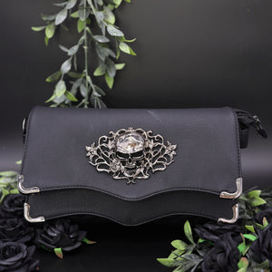 The GothX Skulls and Roses Quilted Clutch Bag sat on a black background with green foliage and black roses surrounding it. The vegan leather clutch bag is facing forward to highlight the two magnetic clip close flaps with metal corners, a stitch quilted front, a skulls and roses metal centrepiece and two D rings on either side for a detachable strap. The mini bag is inspired by gothic grunge witchy fashion.