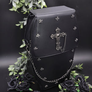 The gothx coffin vegan backpack is sat on a black studio background surrounded by black roses and green foliage. The bag is facing forward to highlight the cross mini studs, cross and chain emblem and detachable silver chain.