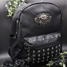 Load image into Gallery viewer, The GothX Skulls and Roses Vegan Backpack on a black studio background with black roses and foliage surrounding it. The black backpack has silver skull and cross studs along the front zip pocket and a rose skull emblem in the middle.
