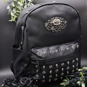 The GothX Skulls and Roses Vegan Backpack on a black studio background with black roses and foliage surrounding it. The black backpack has silver skull and cross studs along the front zip pocket and a rose skull emblem in the middle.