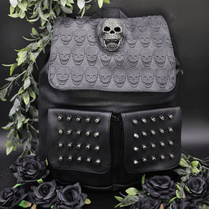 GothX twin pocket skull vegan backpack on a black background with leaves surrounding it. The bag is facing forward to highlight the diamante effect skull, skull embossed vegan leather front flap, tassel tie cords and two silver studded front pockets.