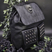 Load image into Gallery viewer, GothX twin pocket skull vegan backpack on a black background with leaves surrounding it. The bag is facing forward angled to the right to highlight the diamante effect skull, skull embossed vegan leather front flap, tassel tie cords and two silver studded front pockets.
