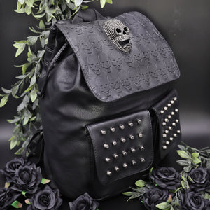 GothX twin pocket skull vegan backpack on a black background with leaves surrounding it. The bag is facing forward angled to the right to highlight the diamante effect skull, skull embossed vegan leather front flap, tassel tie cords and two silver studded front pockets.
