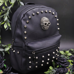 The GothX Skull Head Small Studded Vegan Mini Backpack on a black studio background with black roses and green vines surrounding them. Bag is facing forward angled right to highlight the diamanté style skull, gunmetal coloured studs and front zip pocket.