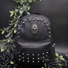 Load image into Gallery viewer, The GothX Skull Head Small Studded Vegan Mini Backpack on a black studio background with black roses and green vines surrounding them. The vegan leather bag is facing forward to highlight the diamanté style skull, gunmetal coloured studs and front zip pocket.
