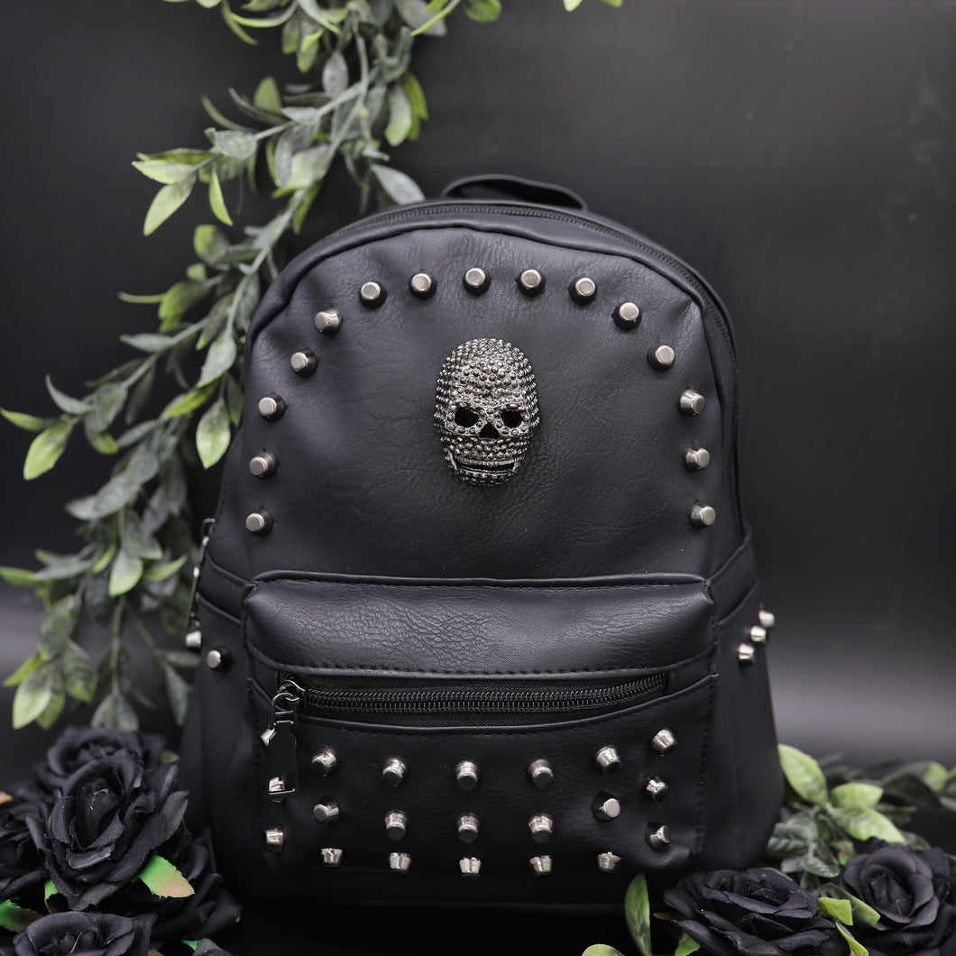 The GothX Skull Head Small Studded Vegan Mini Backpack on a black studio background with black roses and green vines surrounding them. The vegan leather bag is facing forward to highlight the diamanté style skull, gunmetal coloured studs and front zip pocket.