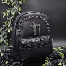 Load image into Gallery viewer, The GothX black cross vegan stud mini backpack on a black background with black roses and green leaf foliage surrounding it. The bag is facing forwards angled to the right to highlight the cross studs along the top zip line and top of zipped pocket, studded cross with chain centrepiece and side studs on the slot pockets.
