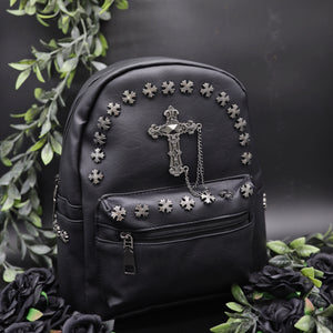The GothX black cross vegan stud mini backpack on a black background with black roses and green leaf foliage surrounding it. The bag is facing forwards angled to the right to highlight the cross studs along the top zip line and top of zipped pocket, studded cross with chain centrepiece and side studs on the slot pockets.