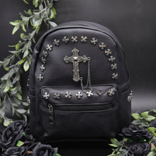 Load image into Gallery viewer, The GothX black cross vegan stud mini backpack on a black background with black roses and green leaf foliage surrounding it. The bag is facing forwards to highlight the cross studs along the top zip line and top of zipped pocket, studded cross with chain centrepiece and side studs on the slot pockets.
