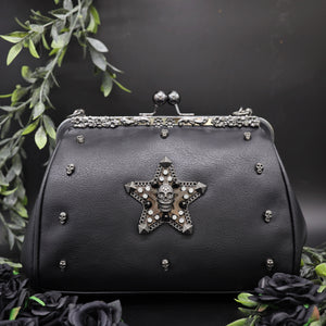 The GothX Skull-Struck vegan vintage clasp handbag on a black background with black roses and foliage surrounding it. The bag is facing forward to highlight the vintage ball clasp close, floral metal top detailing, detachable handle strap, skull studs and skull star with chains, studs and crystals.