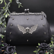 Load image into Gallery viewer, The GothX winged skull vegan vintage clasp handbag on a black studio background with black roses and green foliage surrounding it. The bag is facing forward to highlight the ball clasp close, metal floral detailing along the top, mini skull studs and winged skull centre piece. 
