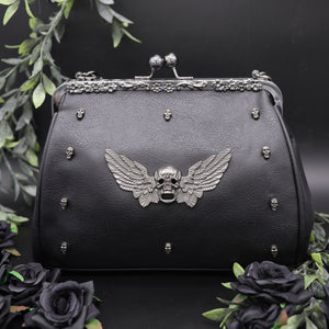 The GothX winged skull vegan vintage clasp handbag on a black studio background with black roses and green foliage surrounding it. The bag is facing forward to highlight the ball clasp close, metal floral detailing along the top, mini skull studs and winged skull centre piece. 