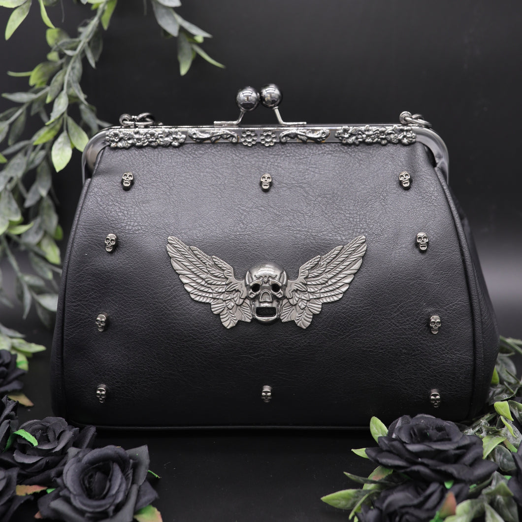 The GothX winged skull vegan vintage clasp handbag on a black studio background with black roses and green foliage surrounding it. The bag is facing forward to highlight the ball clasp close, metal floral detailing along the top, mini skull studs and winged skull centre piece. 