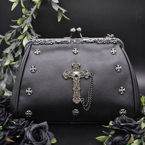 The GothX Don't Cross Me Vegan Vintage Clasp Handbag on a black studio background with black roses and green foliage surrounding it. The bag is facing forward to highlight the floral metal detailing along the top, vintage ball clasp close, mini cross studs and large cross chain emblem.